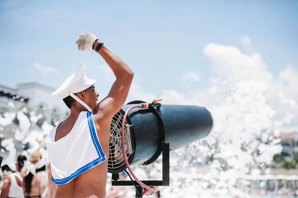 Beach-goer in white swimwear and hat holding binoculars with ocean crowd and boats in background
