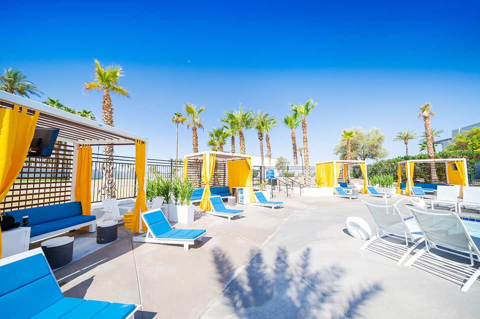 Poolside cabanas with yellow curtains, blue loungers, and palm tree landscaping