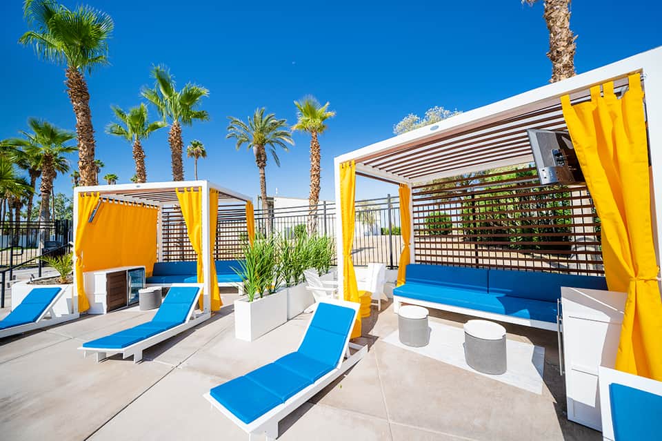 Modern pool cabanas with yellow curtains, blue loungers, and palm trees under blue sky