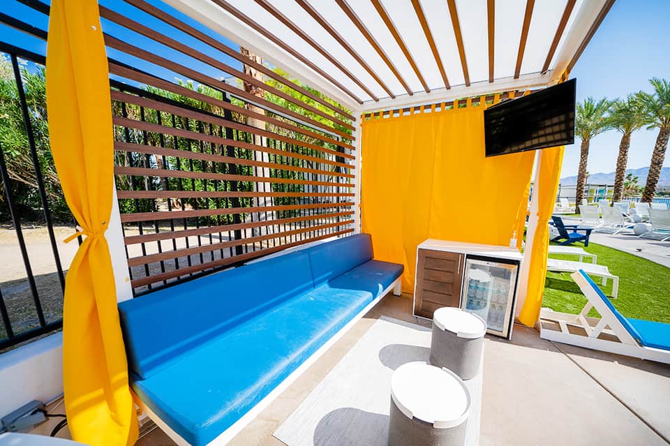 Modern pool cabana with bright yellow curtains, blue seating, and wooden lattice design