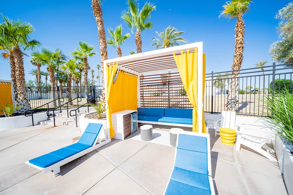 Colorful poolside cabana with yellow curtains, blue seating, and palm trees in desert setting