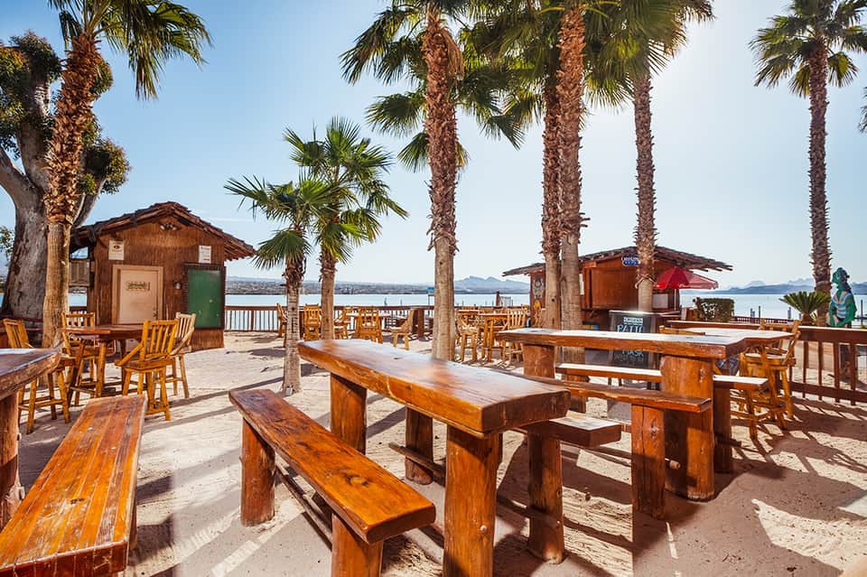 Beachfront bar with wooden tables, palm trees, and water views with mountain backdrop