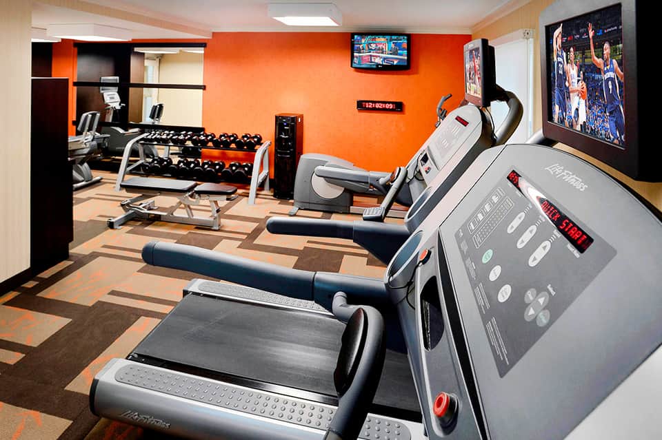 Hotel fitness center with treadmills, dumbbells, exercise bikes, orange accent wall, and sports TV display.