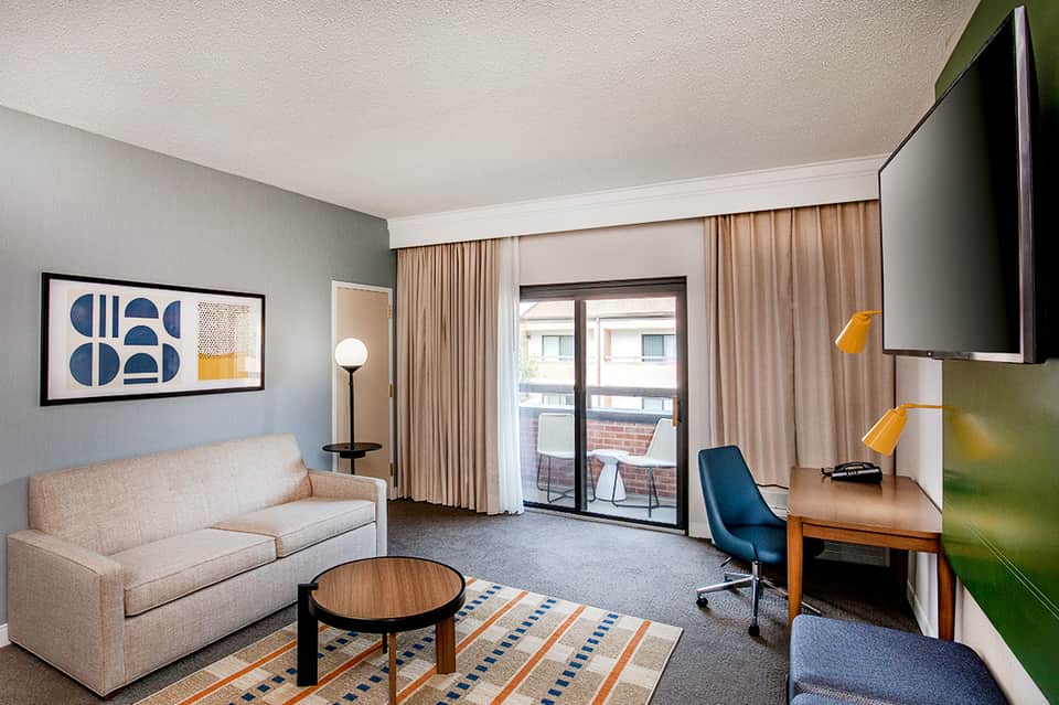 Modern hotel suite with beige sofa, desk, blue accent chair, and balcony