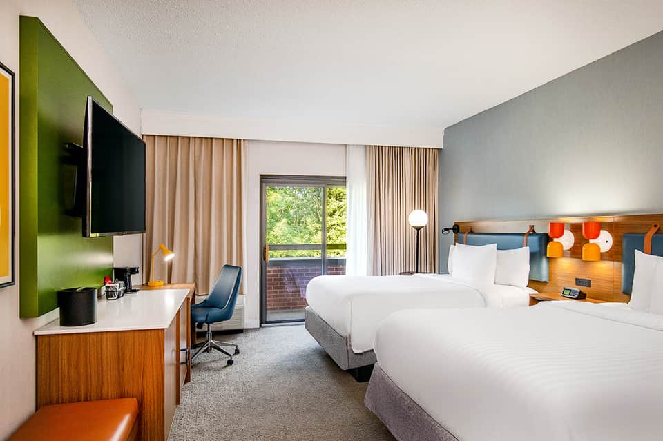 Contemporary hotel room with two queen beds, green accent wall, wood headboard, and balcony access