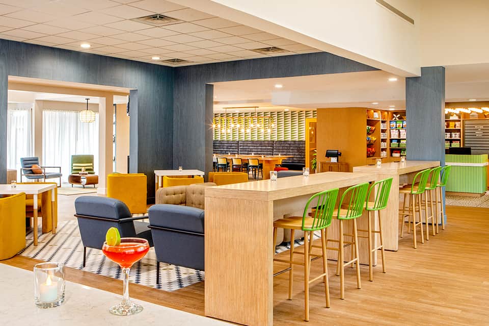 Contemporary hotel lobby bar with orange and green accents, wooden furnishings, and open layout