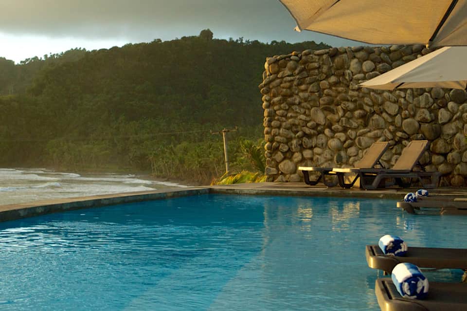 Luxury resort pool with river view, stone wall feature, loungers, and tropical vegetation