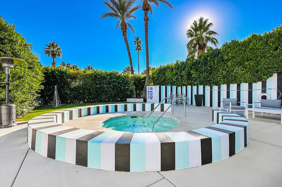 Colorful striped spa pool surrounded by white cabanas, hedges, palm trees, and sunny courtyard