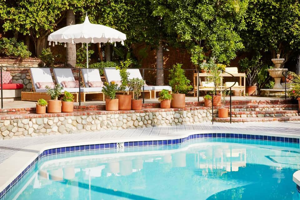 Luxury resort pool with white umbrella, lounge chairs with pink cushions, landscaped stone wall, and mature greenery backdrop