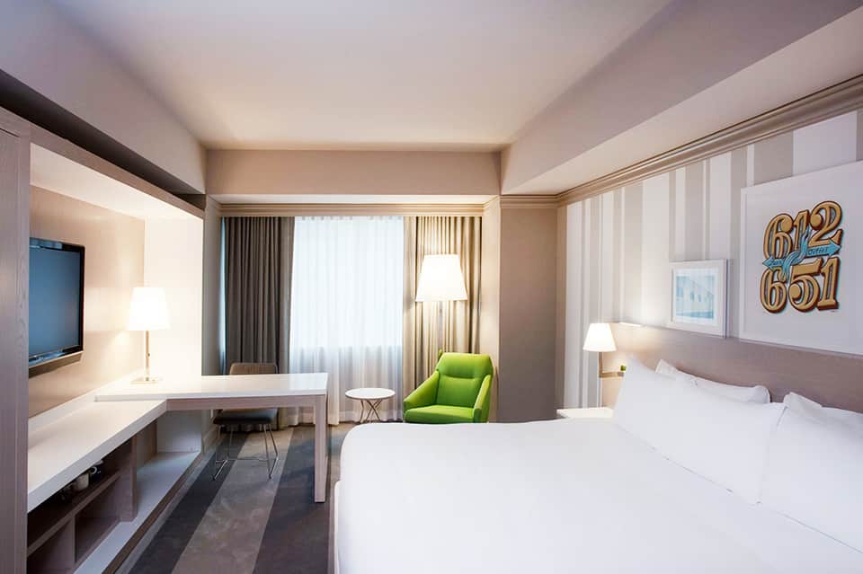 Contemporary hotel room with king bed, work desk, green accent chair, and floor-to-ceiling windows