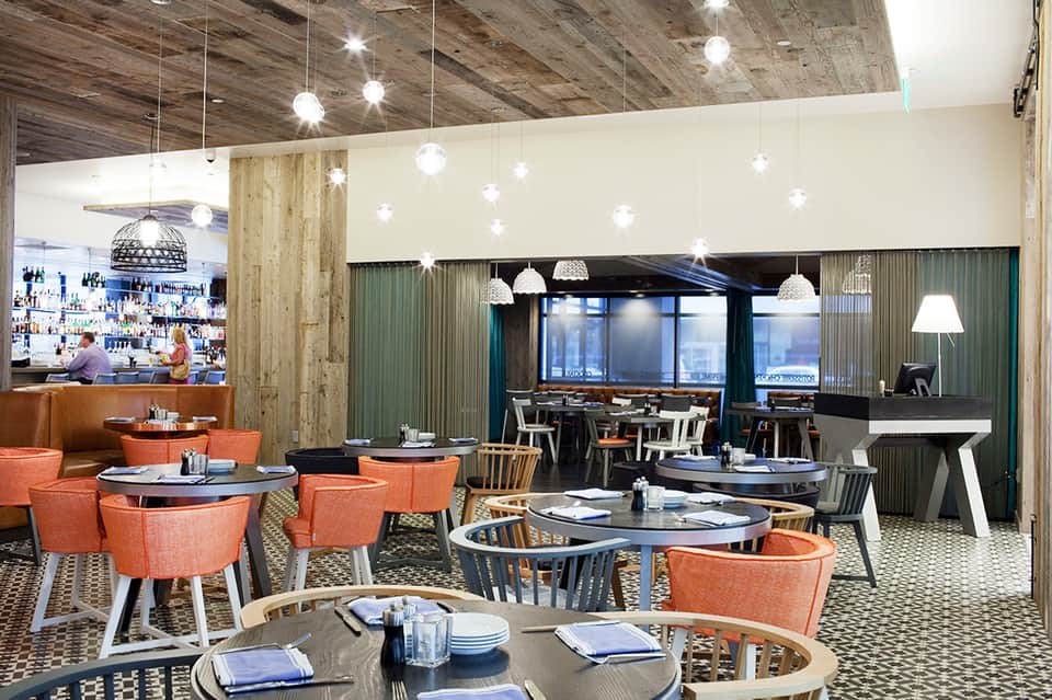 Modern restaurant interior with orange chairs, hanging lights, wooden beams, and ocean views
