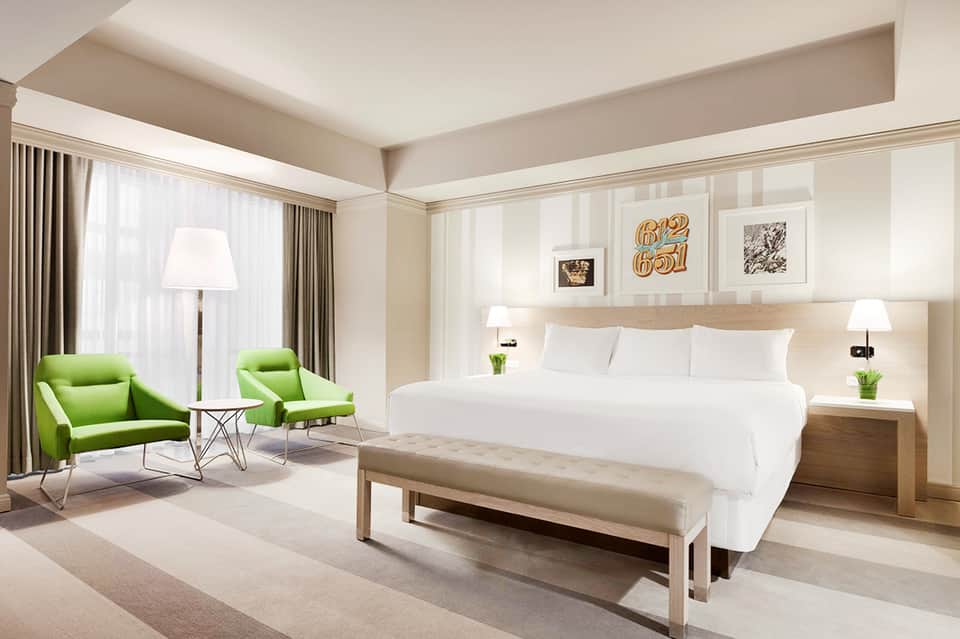 Luxury hotel suite with king bed, green lounge chairs, floor-to-ceiling windows, and contemporary light wood furniture