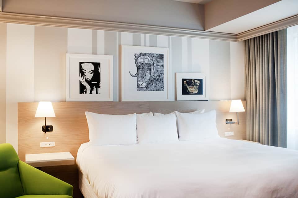 Modern hotel bedroom with white bedding, wood headboard accent wall, framed artwork, and green chair