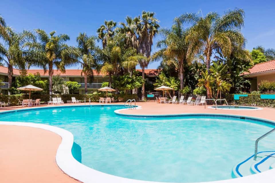 Resort pool with spa area, palm trees, umbrellas, and tropical landscaping under clear sky