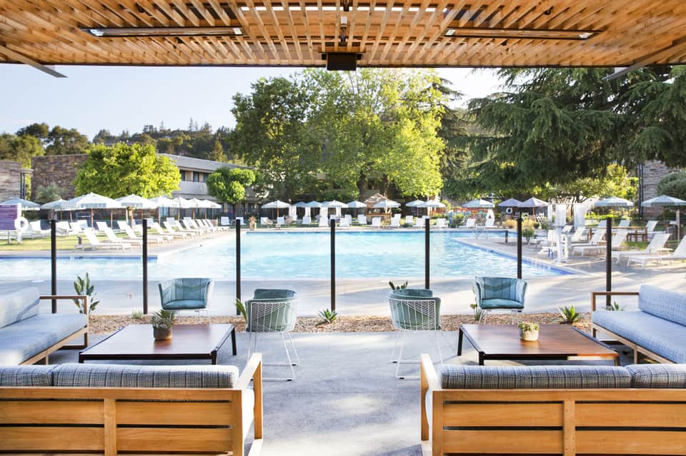 Daytime resort pool scene with wooden pergola, loungers with blue cushions, umbrellas, and trees