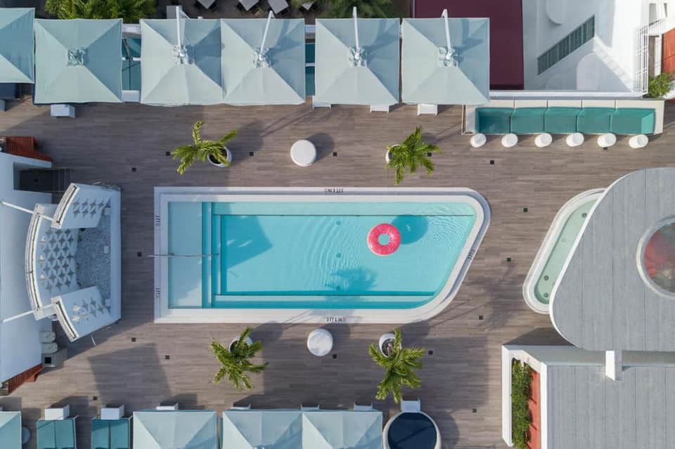 Overhead view of modern resort pool surrounded by white umbrellas, lounge areas, palm trees, and wooden decking