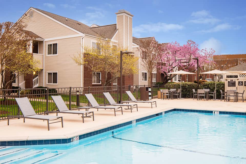 Resort pool area with white lounge chairs, colonial-style brick building, and blooming pink trees