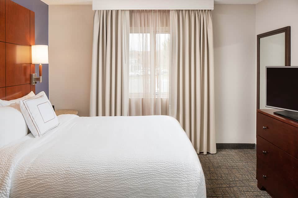 Hotel bedroom with white bedding, warm wood headboard, and window with sheer curtains