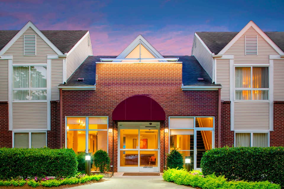 Brick hotel entrance with red awning, white building wings, and manicured green landscaping