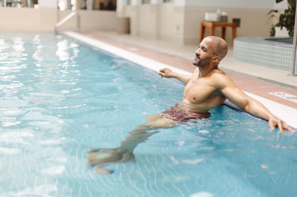 Man relaxing in indoor heated swimming pool with white tiled walls and natural light