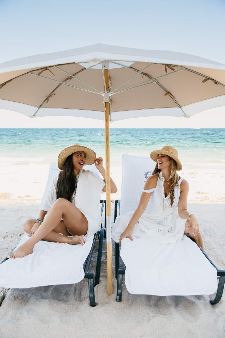 Two women relaxing on beach loungers under large umbrella with ocean and sand background