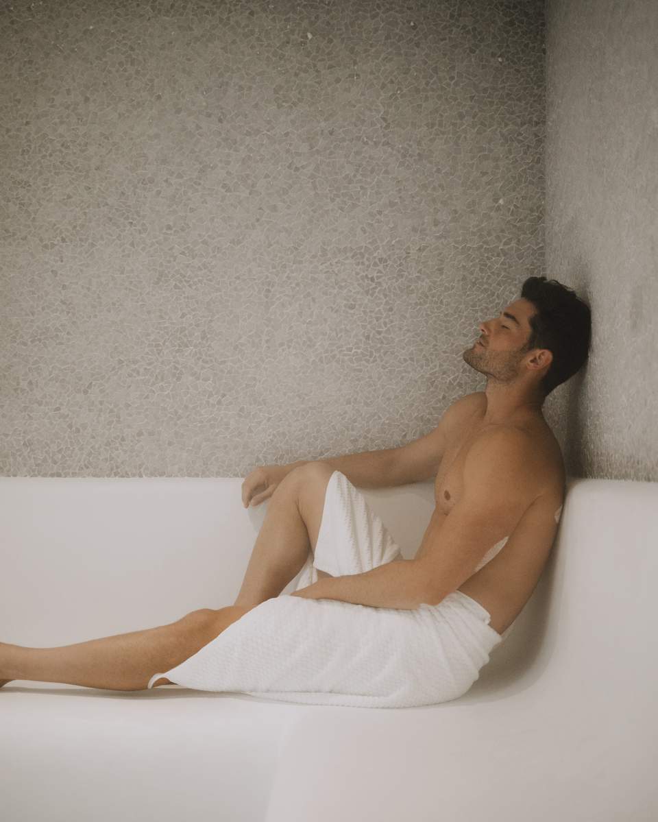Man relaxing in white bathtub wrapped in towel against textured wall