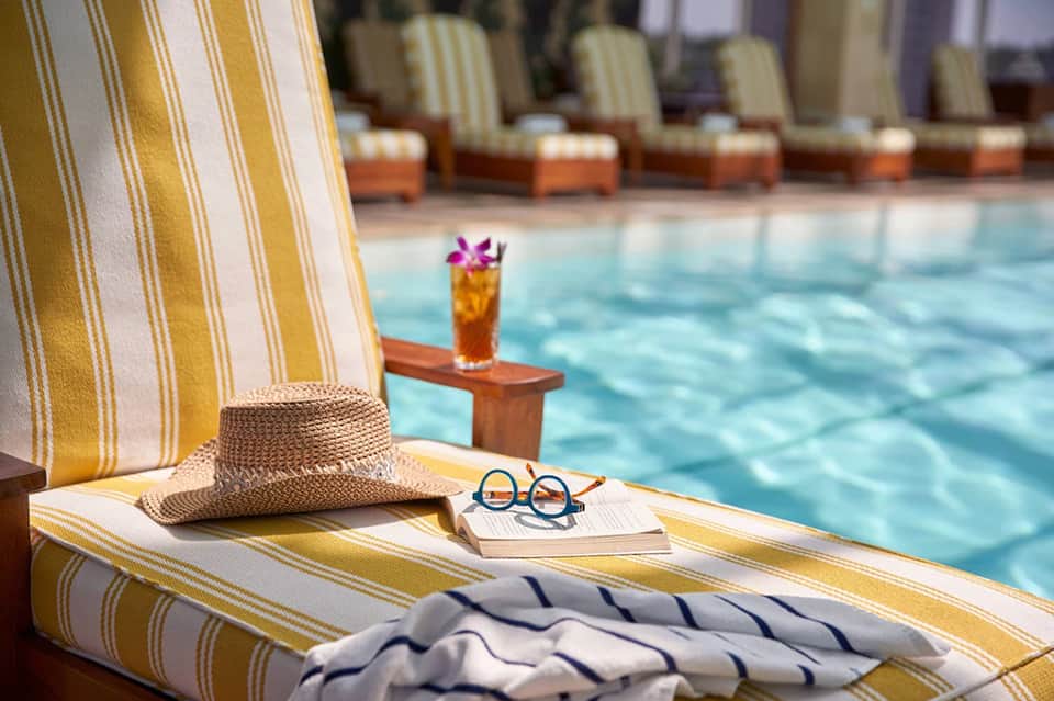 Poolside lounge chair with yellow striped fabric, straw hat, sunglasses, and cocktail with flower garnish