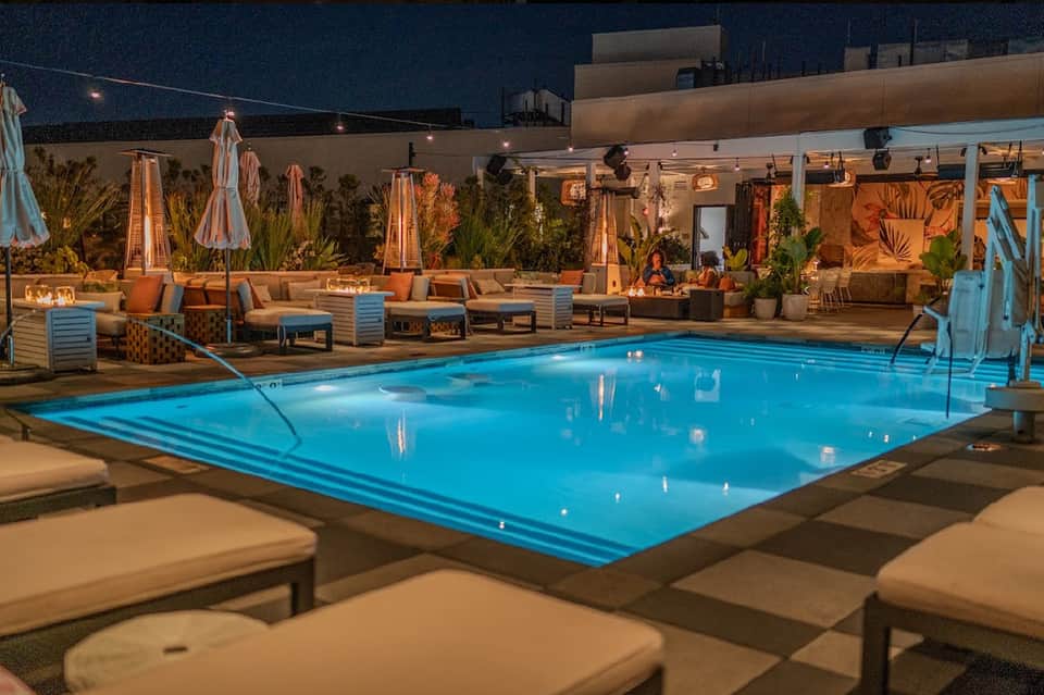 Nighttime resort pool illuminated in blue with fire pits, lounge seating, and striped umbrellas