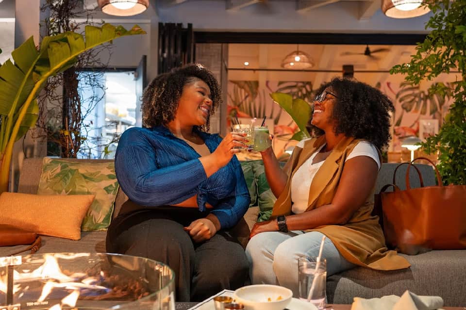 Two women toasting drinks in modern hotel lounge with tropical plants and warm lighting