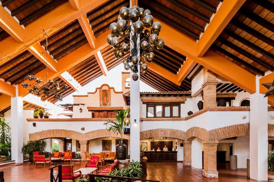 Spanish colonial hacienda-style lobby with orange wooden beams, arched brick doorways, red seating, and decorative chandelier