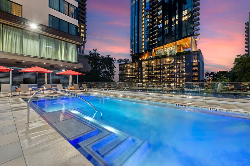 Illuminated rooftop pool at dusk with red umbrellas and city skyline backdrop