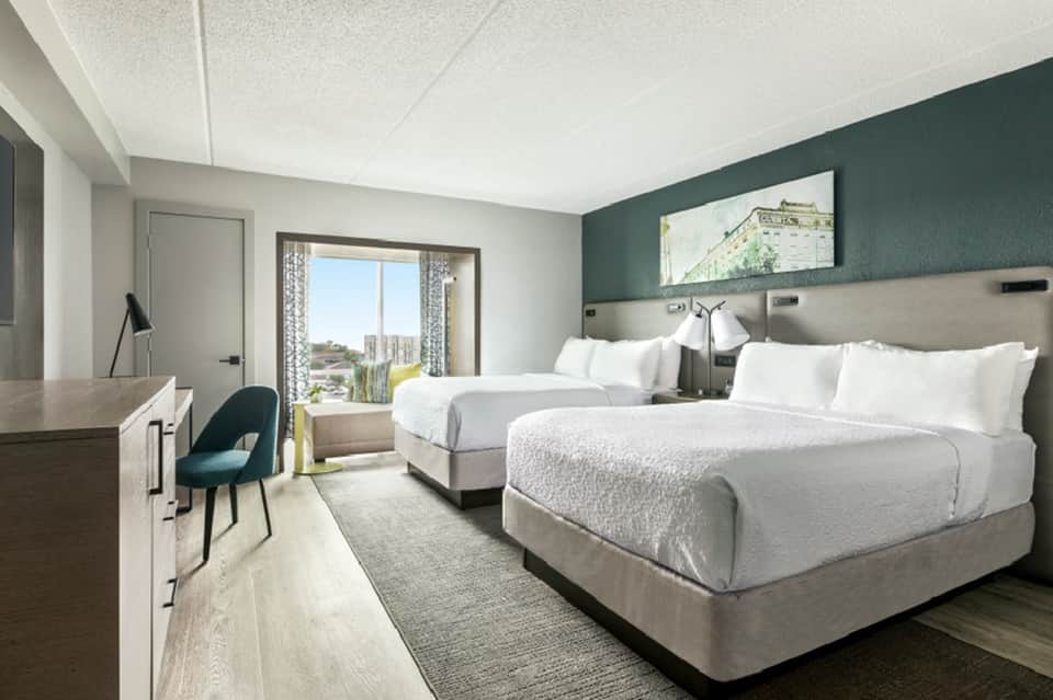 Hotel guest room with two queen beds, teal accent wall, desk, and balcony with city views