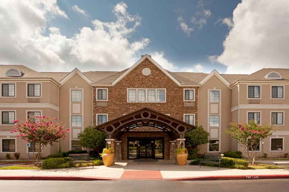 Hotel exterior with tan and brick facade, arched entrance, flowering shrubs, and manicured grounds