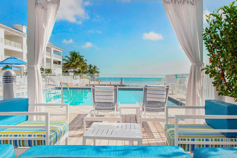Oceanfront balcony view with lounge chairs, pool, palm trees, and white curtains framing the sea