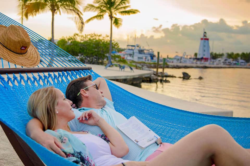 Couple relaxing in hammock overlooking marina with sailboats and lighthouse at sunset