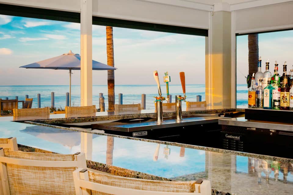 Oceanfront bar with coastal views, palm trees, and blue umbrellas overlooking the sea