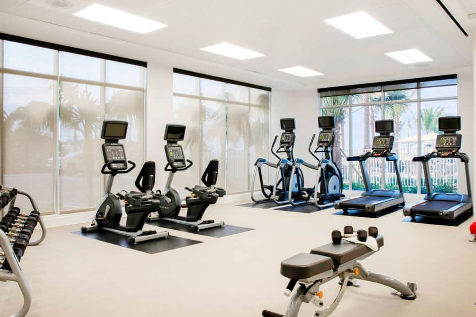 Bright fitness center with cardio equipment, exercise bikes, treadmills, and large windows overlooking palm trees
