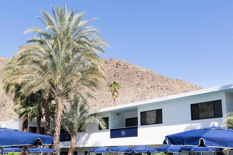 Desert resort building with palm trees and blue umbrellas against mountainous backdrop