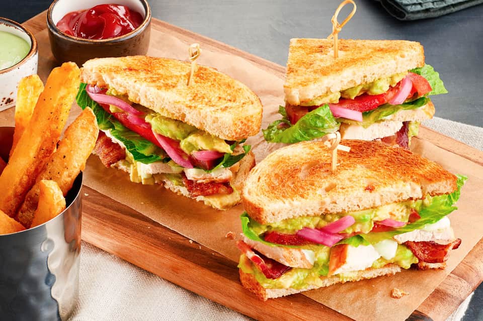 Three gourmet sandwiches with avocado, bacon, and fresh vegetables served with golden fries on a wooden board