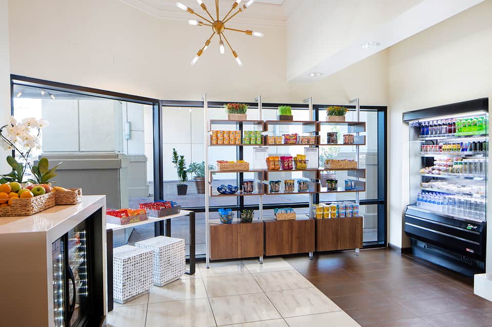 Contemporary hotel cafe with shelving, refrigerated displays, food counter, and potted plants
