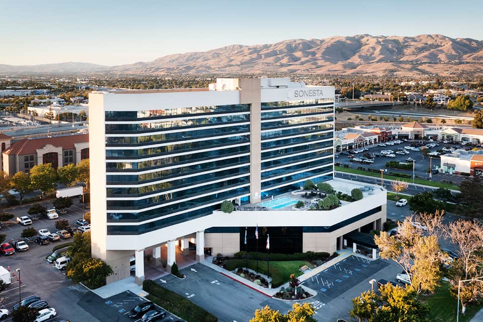 Modern glass hotel building with pool on rooftop, mountains and water visible in background landscape