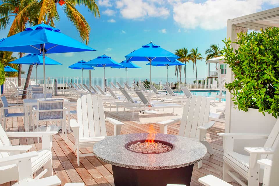Beachfront deck with blue umbrellas, white adirondack chairs, and a fire pit overlooking the ocean