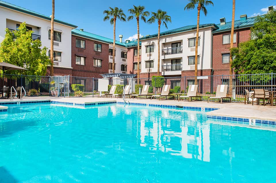 Rectangular swimming pool with turquoise water, multi-story brick hotel buildings, and palm trees