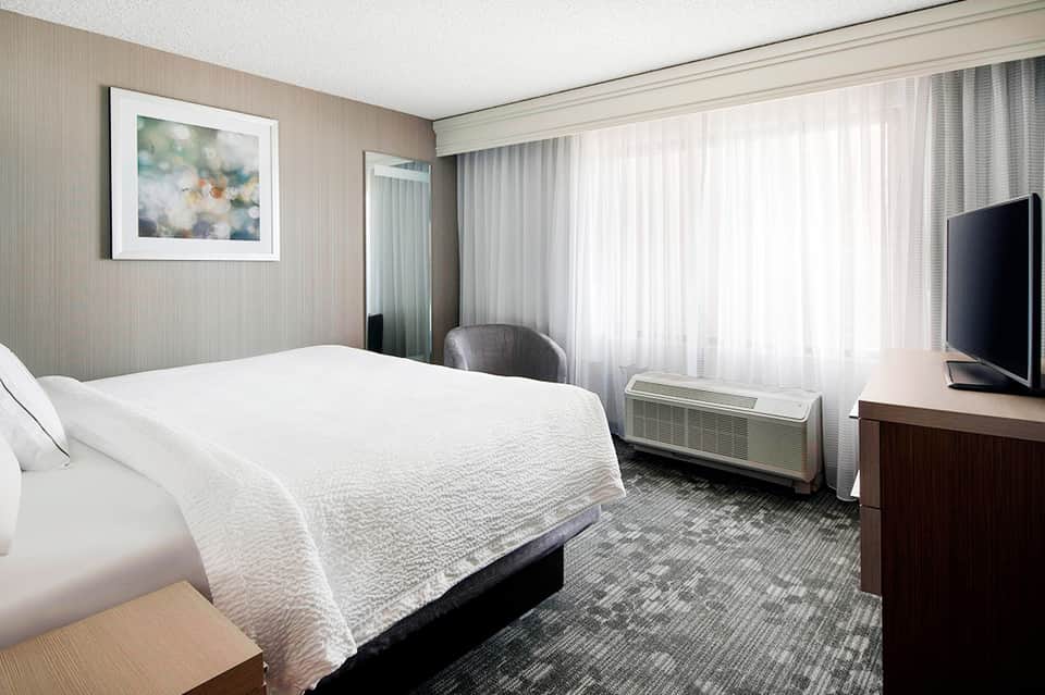 Modern hotel bedroom with white bedding, desk area, and floor-to-ceiling windows