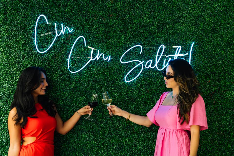 Two women in colorful dresses holding wine glasses in front of a neon 'Gin Salsa' sign on green hedge wall