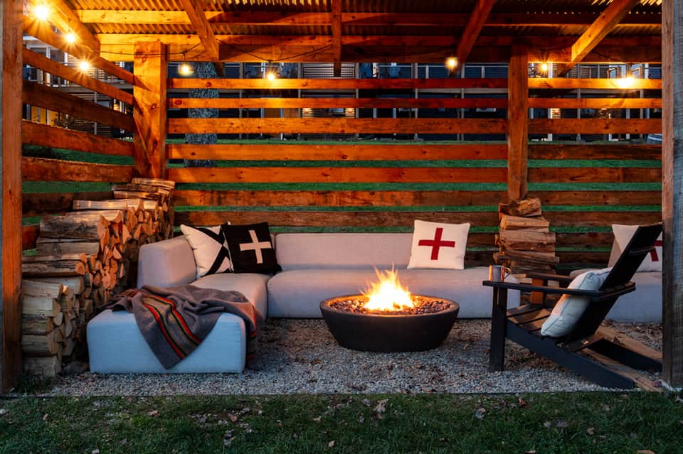 Outdoor fire pit lounge with white seating, rustic wood backdrop, and decorative pillows