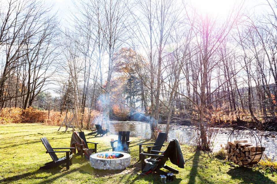 Waterside fire pit with Adirondack chairs surrounded by bare trees and flowing creek