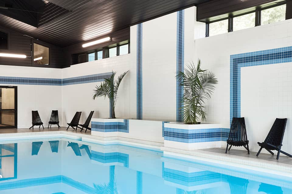 Modern indoor pool with white tile walls, blue accents, and black lounge chairs