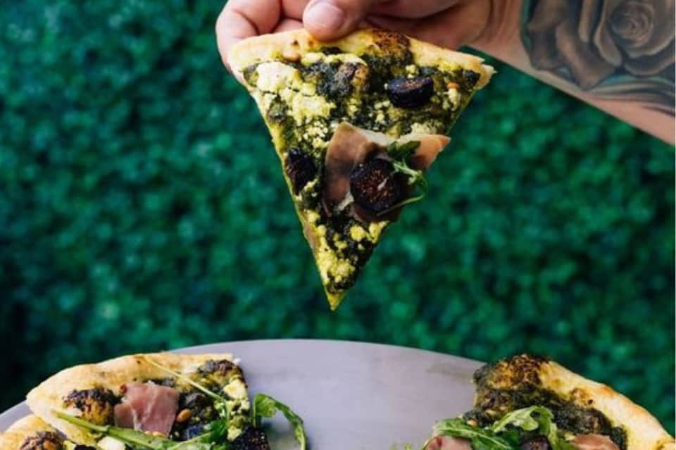 Hand holding fresh pizza slice with vegetables over table with pesto pizza