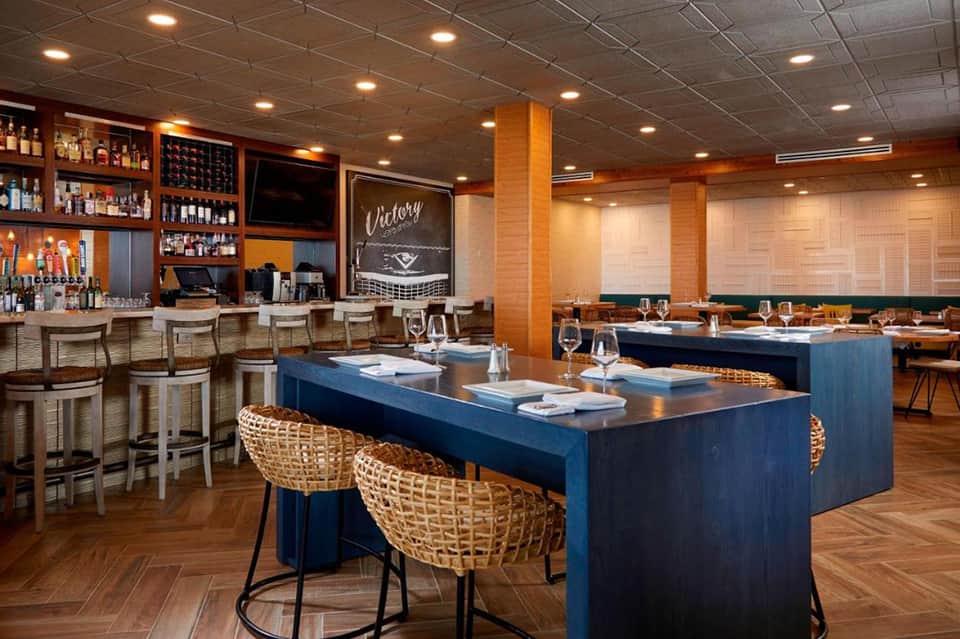 Upscale restaurant interior with blue counters, woven chairs, bar, and warm wood finishes